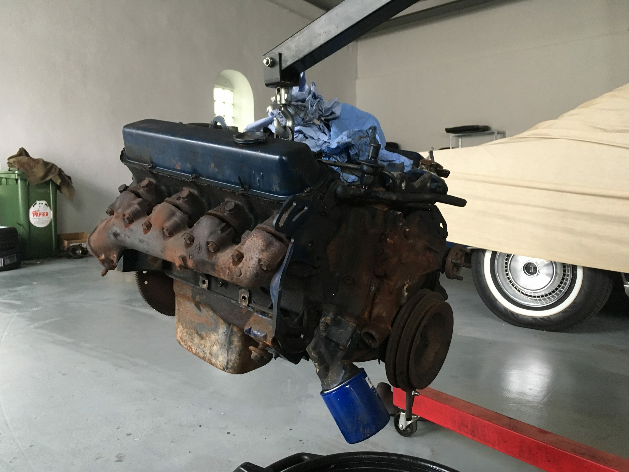 Rostiger alter Motor eines klassischen US-Fahrzeugs (z.B. Cadillac/Lincoln) wird in der Werkstatt von Oldtimer Profi gehoben – Vorbereitung für die fachgerechte Reparatur oder Instandsetzung.