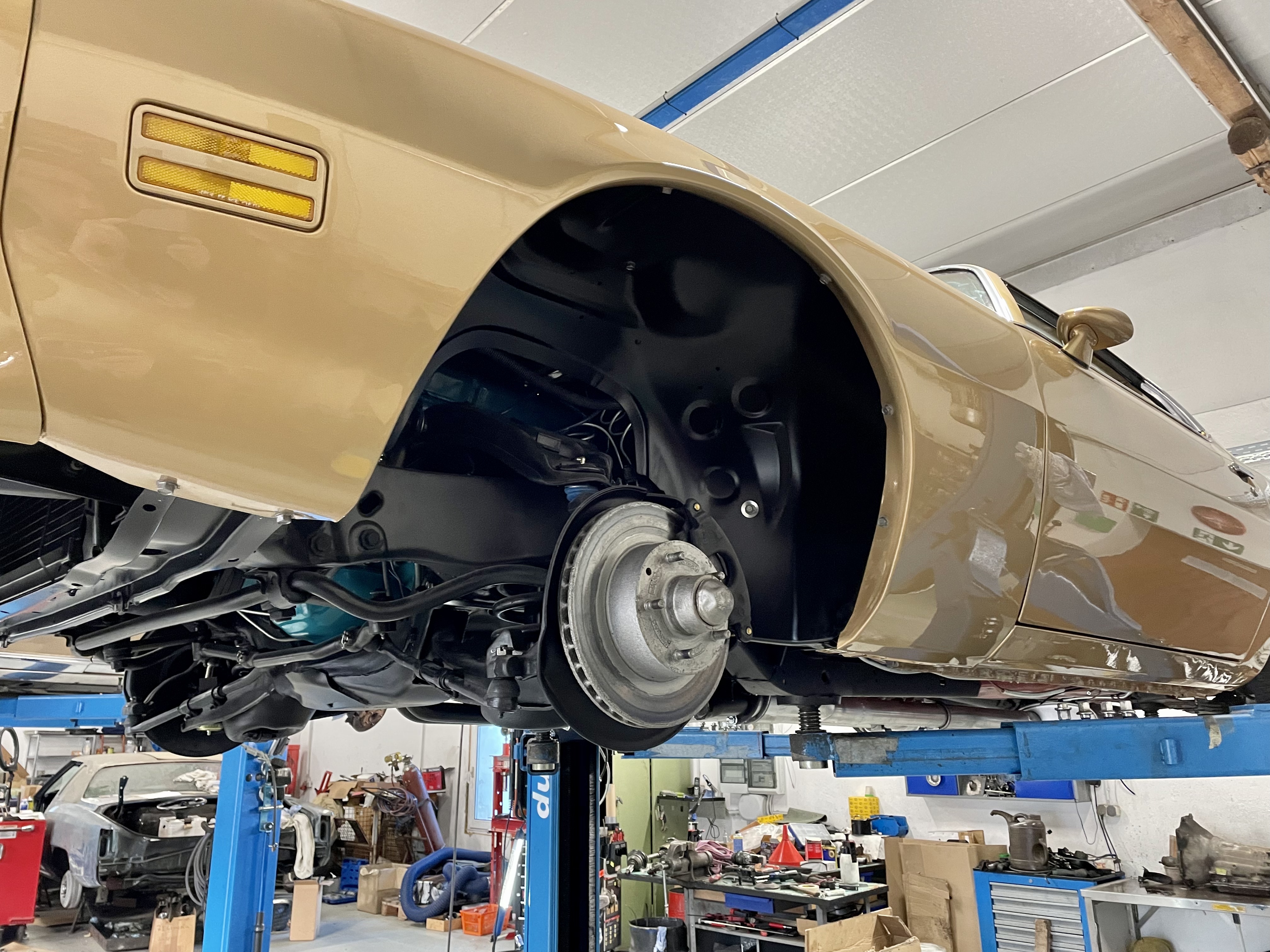 Beige Oldtimer auf blauer Hebebühne in einer Werkstatt, Rad und Unterboden sichtbar – spezialisierte Reparatur und Pflege klassischer US-Fahrzeuge wie Cadillac und Lincoln (Baujahre 1956-1979) bei Oldtimer Profi.