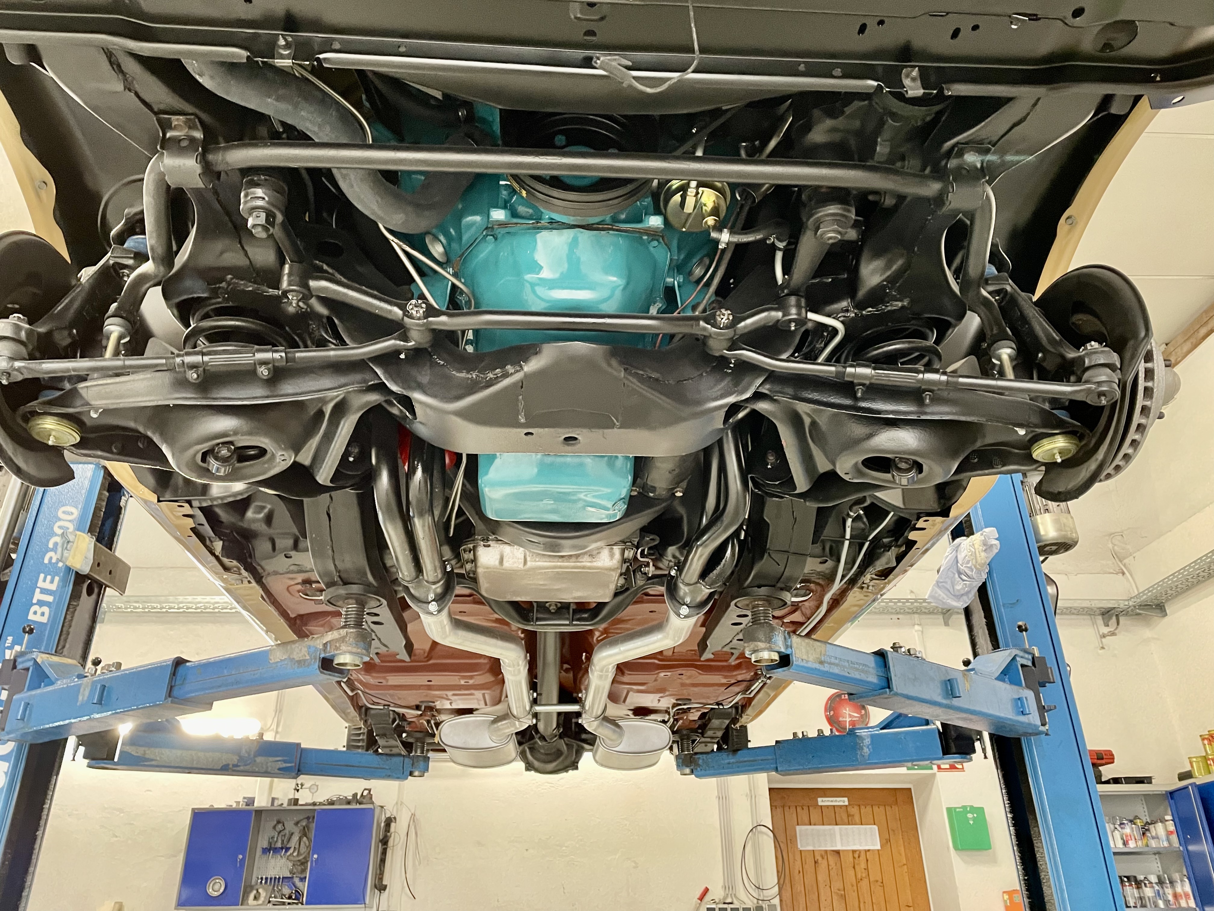 Unterseite eines klassischen US-Oldtimers auf einer Hebebühne, mit sichtbarem türkisfarbenem Motor und Fahrwerk, in der spezialisierten Werkstatt für Cadillac und Lincoln (Oldtimer Profi).