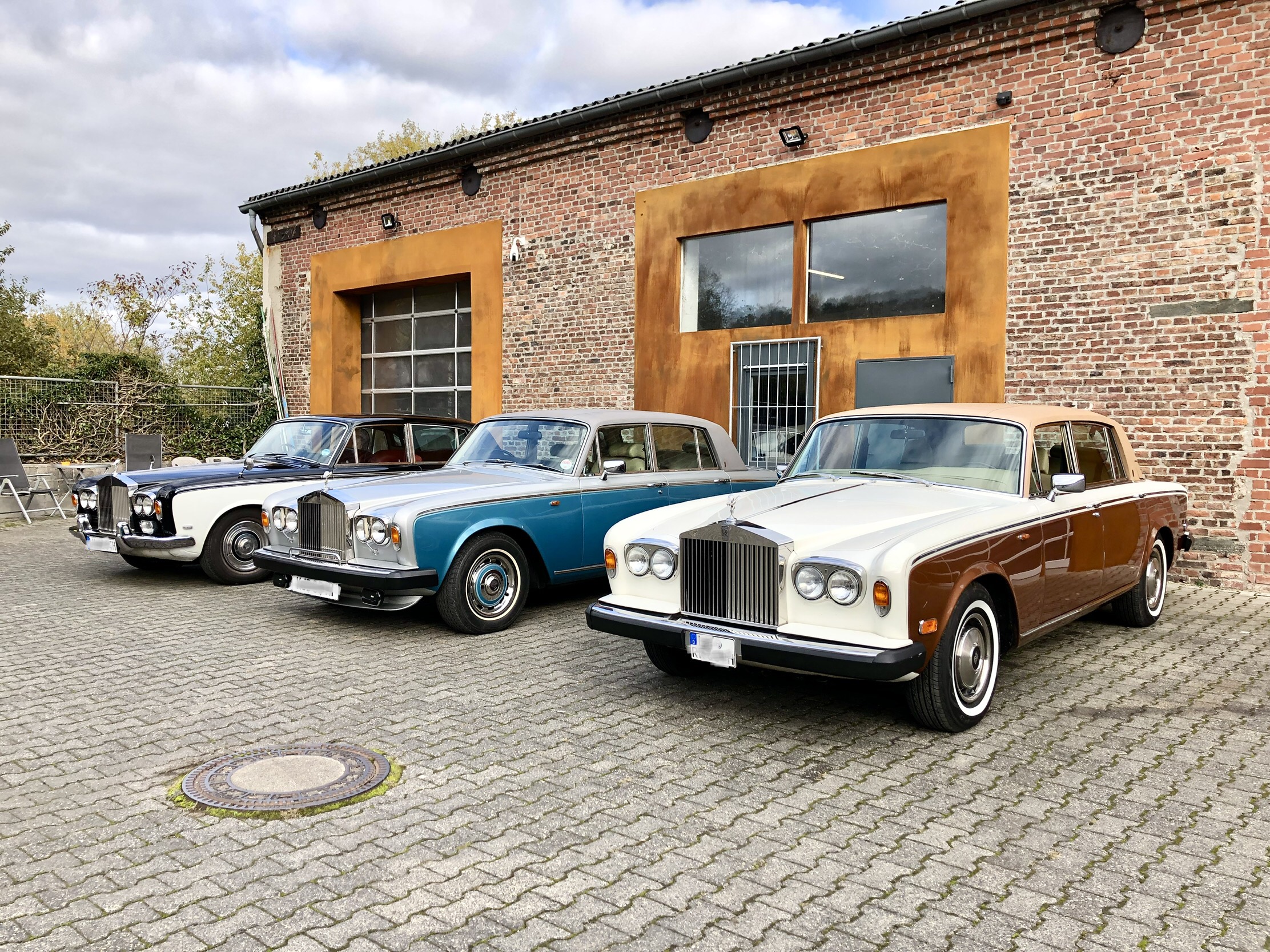 Drei klassische Luxusautos stehen auf einem gepflasterten Hof vor einem Backsteingebäude – repräsentativ für die Fachwerkstatt Oldtimer Profi, die sich auf die Reparatur und Pflege von klassischen US-Fahrzeugen (z. B. Cadillac, Lincoln) spezialisiert.