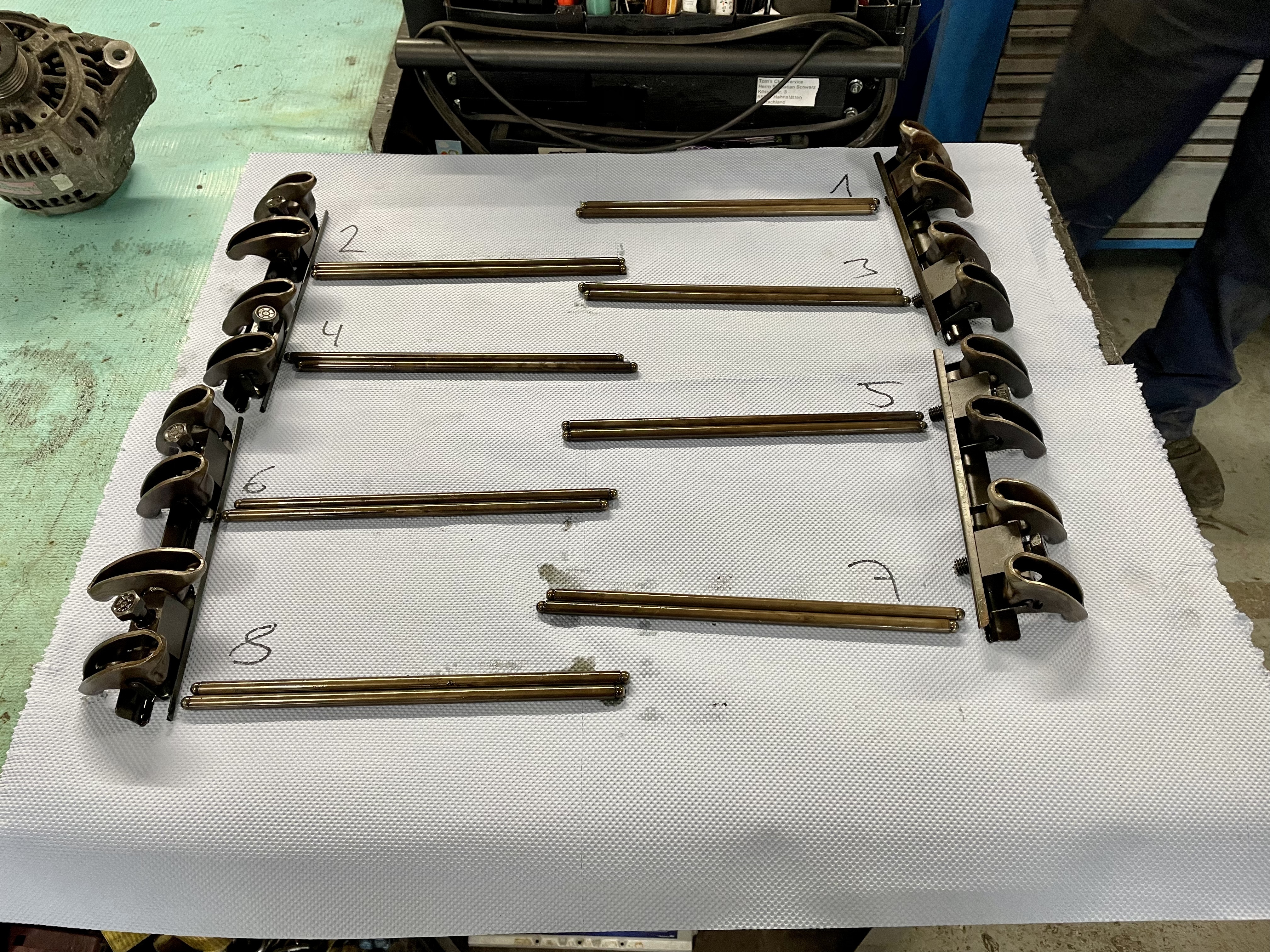 Disassemblierte Ventiltriebskomponenten (Kipphebel, Stößelstangen) eines klassischen US-Automotors, nummeriert und auf einem weißen Arbeitsunterlagen sortiert, in der Werkstatt von Oldtimer Profi – spezialisiert auf Cadillac und Lincoln 1956-1979.